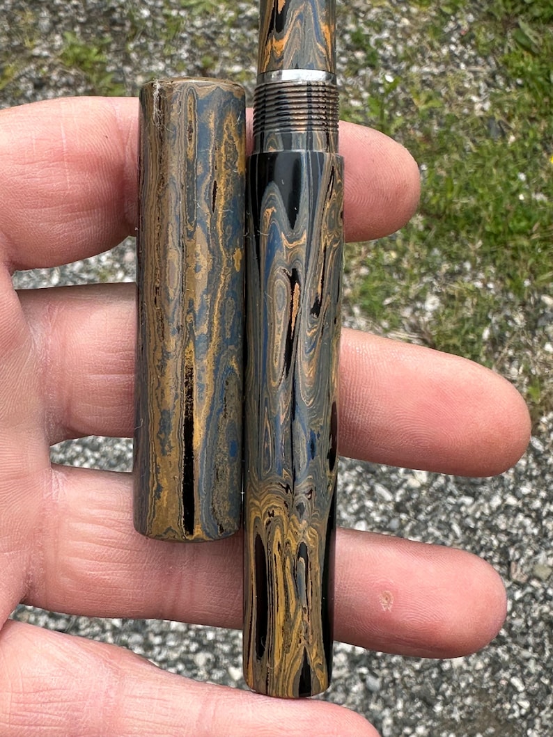 May include: A handcrafted pen and pen case with a unique swirling pattern in shades of black, gold, and blue. The pen features a silver metal band near the nib. The pen case is cylindrical and matches the pen's design.