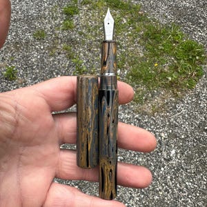 May include: A fountain pen with a marbled pattern in shades of black, gold, and blue. The pen's nib is silver, and the cap is detached, revealing the pen's design. The pen is held in a hand against a background of gravel and grass.
