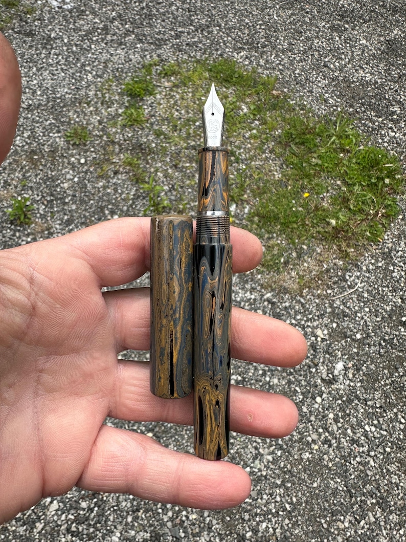 May include: A fountain pen with a marbled pattern in shades of black, gold, and blue. The pen's nib is silver, and the cap is detached, revealing the pen's design. The pen is held in a hand against a background of gravel and grass.