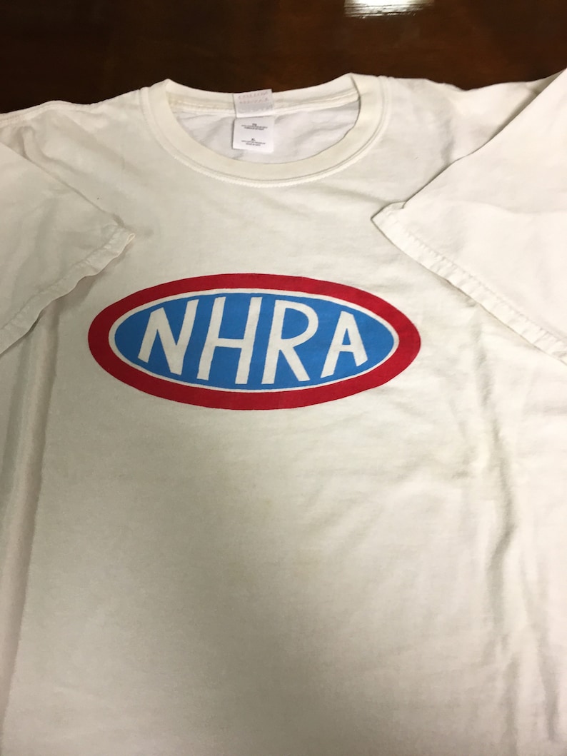 May include: White t-shirt with a red and blue oval logo that says "NHRA" in blue letters.