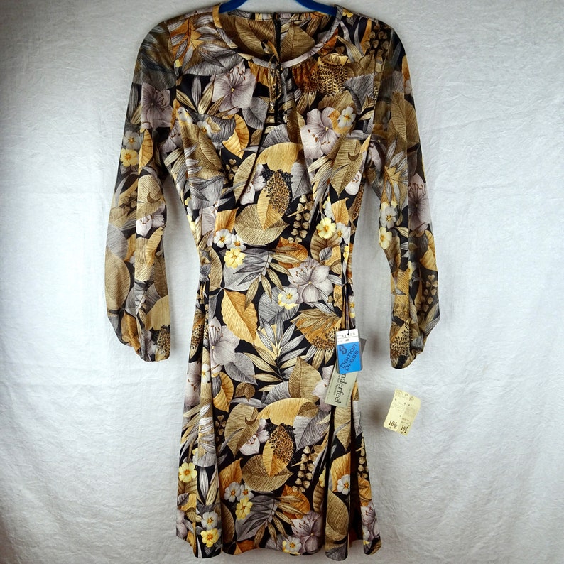 May include: A vintage dress with a floral pattern featuring large leaves and flowers in shades of brown, tan, gray, and yellow on a black background. The dress has long sleeves and a tie at the waist. A tag is attached.