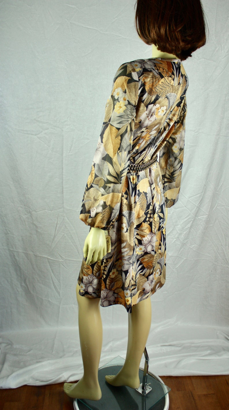 May include: A vintage dress with a floral pattern in shades of black, gold, and cream. The dress features long sleeves, a cinched waist with a gold chain belt, and a knee-length hemline. The dress is displayed on a mannequin.