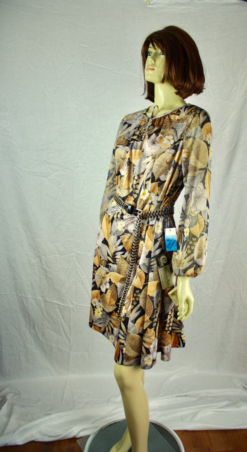 May include: A long-sleeved dress with a floral pattern in shades of brown, beige, and black. The dress features a tie neckline and a chain belt. The dress is displayed on a mannequin. The tag reads "Damce Dress".