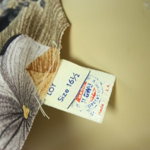 May include: Close-up of fabric with a floral pattern in shades of gray, brown, and black. A tag with the text "Size 16 1/2" and a stamp is attached to the fabric. The fabric is likely part of a garment.
