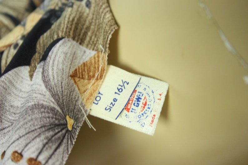 May include: Close-up of fabric with a floral pattern in shades of gray, brown, and black. A tag with the text "Size 16 1/2" and a stamp is attached to the fabric. The fabric is likely part of a garment.