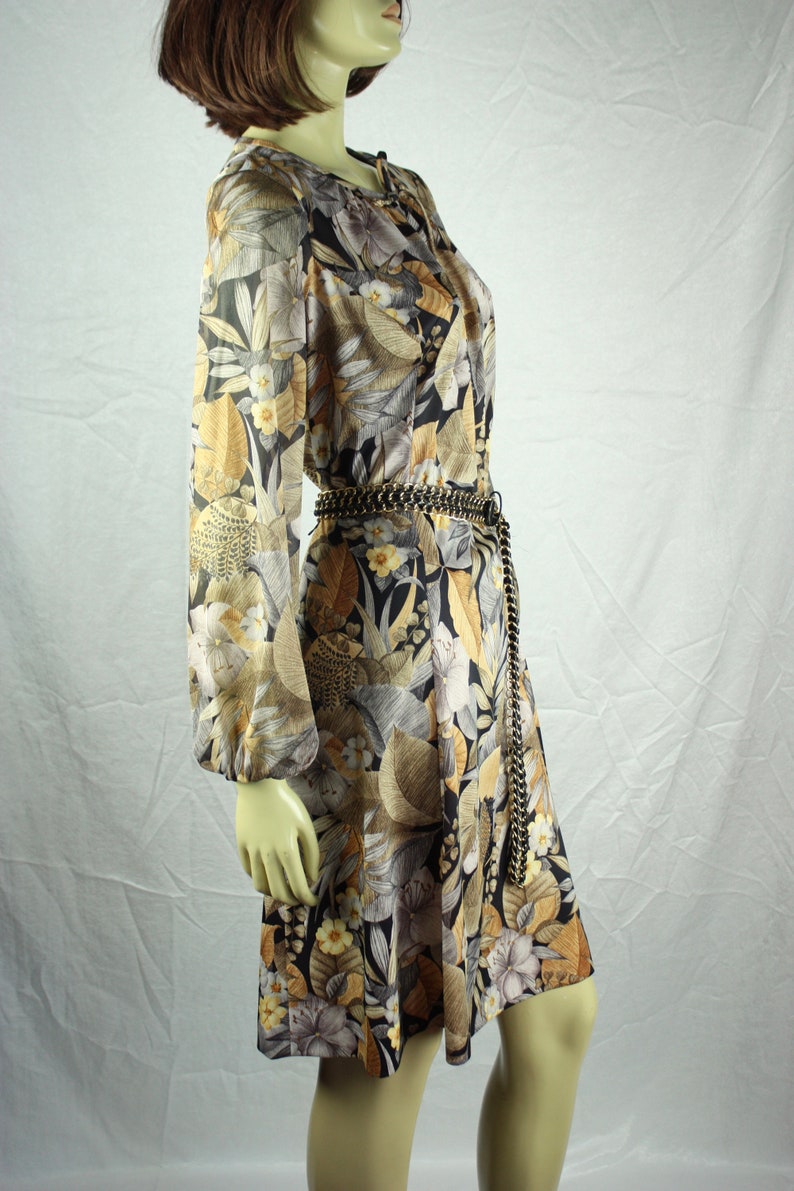 May include: A vintage-style dress with a floral pattern in shades of gold, black, and gray. The dress features long sleeves, a tie-neck detail, and a chain belt. The dress is knee-length.