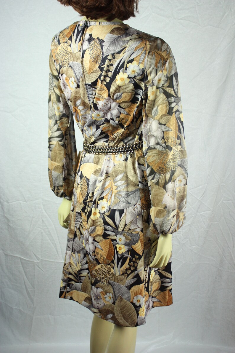 May include: A vintage dress with a botanical print in shades of brown, beige, and black. The long-sleeved dress features a floral pattern, a cinched waist with a chain belt, and a knee-length hemline. The dress is displayed on a mannequin.