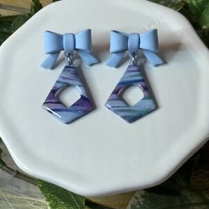 May include: A pair of earrings with a light blue bow and a diamond-shaped pendant. The pendant features a marbled design in shades of blue, purple, and white. The earrings are displayed on a white plate, with greenery in the background.