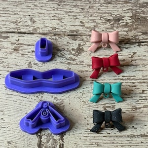 May include: A collection of purple plastic cookie cutters in various shapes, including a bow, a butterfly, and a small cylinder. Several finished bows in pink, red, teal, and black are also displayed.