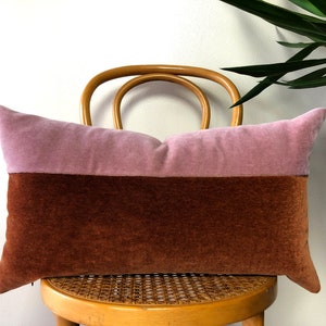 May include: A long rectangular pillow with a two-tone design. The top half is a light dusty rose color and the bottom half is a rich brown color. The pillow is sitting on a wooden chair with a woven seat.