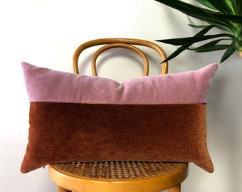 Puede incluir: Una almohada rectangular larga con un diseño de dos tonos. La mitad superior es de color rosa polvoriento claro y la mitad inferior es de color marrón intenso. La almohada está sentada en una silla de madera con un asiento tejido.