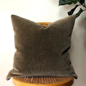 May include: A brown velvet pillow with a subtle texture sits on a woven chair. The pillow is square and has a simple design.
