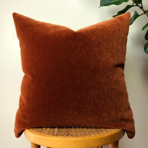 May include: A rust-colored velvet pillow with a square shape and a soft, plush texture. The pillow is sitting on a wooden stool with a woven seat.