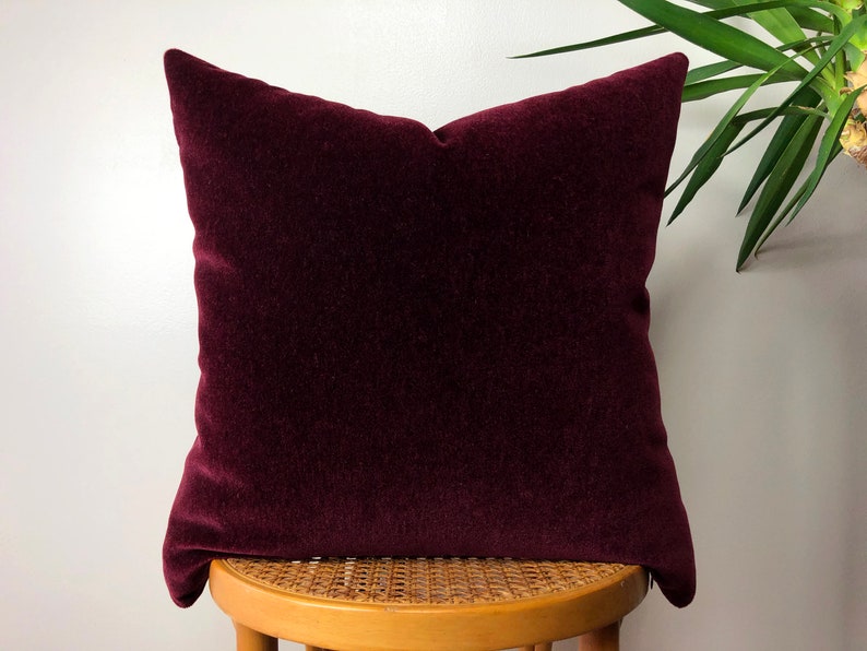 May include: A deep burgundy velvet pillow with a square shape. The pillow is sitting on a wooden stool with a woven seat.