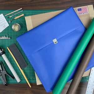 May include: A flat lay of craft supplies for wrapping gifts, including blue, green, and brown wrapping paper, a ruler, a stapler, a brush, and a small jar of gold tacks.