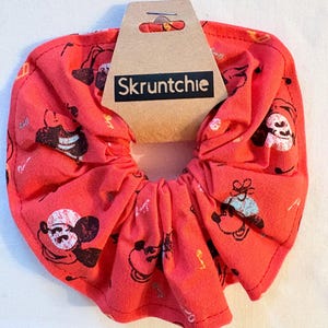 May include: A red scrunchie featuring a repeating pattern of a cartoon character in black and white. The scrunchie has a tag with the text "Skruntchie" in black on a white background.
