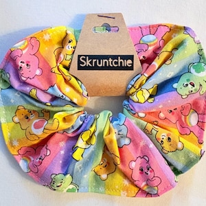 May include: A vibrant scrunchie with a repeating Care Bears design set against a rainbow backdrop. The fabric scrunchie has the word "Skruntchie" printed on a brown tag.