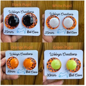May include: Four pairs of round earrings with different colored pupils: black, white, orange, and green. Each earring has an orange and black bat-themed border. The cards read "Wolvyn Creations", "Bat Cave", and sizes 30mm and 35mm.