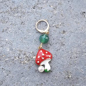 以下が含まれることがあります： 赤と白のキノコのデザインと緑色のガラスビーズが付いた金のチャーム。チャームは金のリングに取り付けられています。
