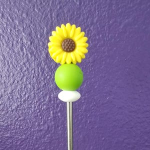 May include: A yellow sunflower with a brown center is attached to a green and white bead on a silver metal stick.