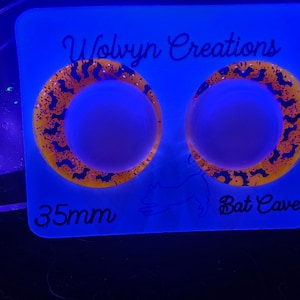May include: Two circular, orange and black earrings with a bat design, displayed on a blue background. The earrings are 35mm in diameter. The text "Wolvyn Creations" and "Bat Cave" are also visible.