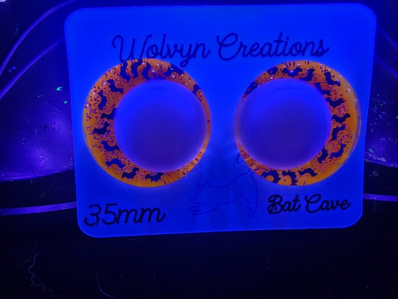 May include: Two circular, orange and black earrings with a bat design, displayed on a blue background. The earrings are 35mm in diameter. The text "Wolvyn Creations" and "Bat Cave" are also visible.