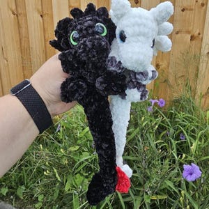 May include: A black and white crochet dragon plushie. The black dragon has green eyes, and the white dragon has blue eyes. The dragons are made of soft, plush yarn and have detailed features. The dragons are shown in a garden setting.