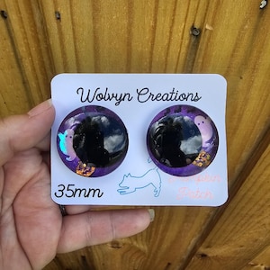 May include: Two round, black and purple earrings with a Halloween theme. Each earring has a black center, surrounded by purple glitter and small ghost and crown designs. The card reads "Wolvyn Creations" and "35mm".