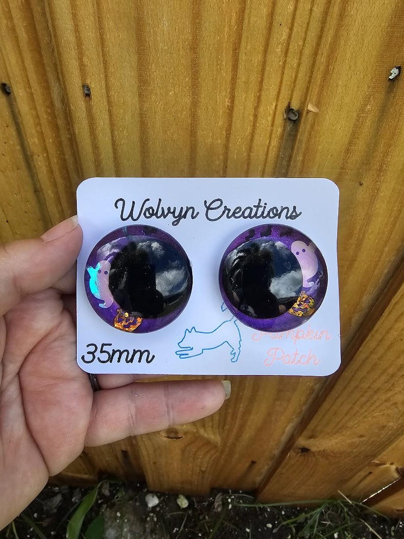 May include: Two round, black and purple earrings with a Halloween theme. Each earring has a black center, surrounded by purple glitter and small ghost and crown designs. The card reads "Wolvyn Creations" and "35mm".