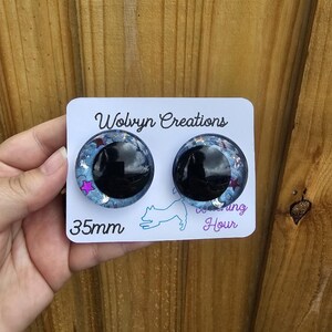 May include: A pair of 35mm round earrings with a black center and a blue, glittery outer ring. The earrings are on a white card with the text "Wolvyn Creations" and a dog silhouette. The earrings are held by a hand.