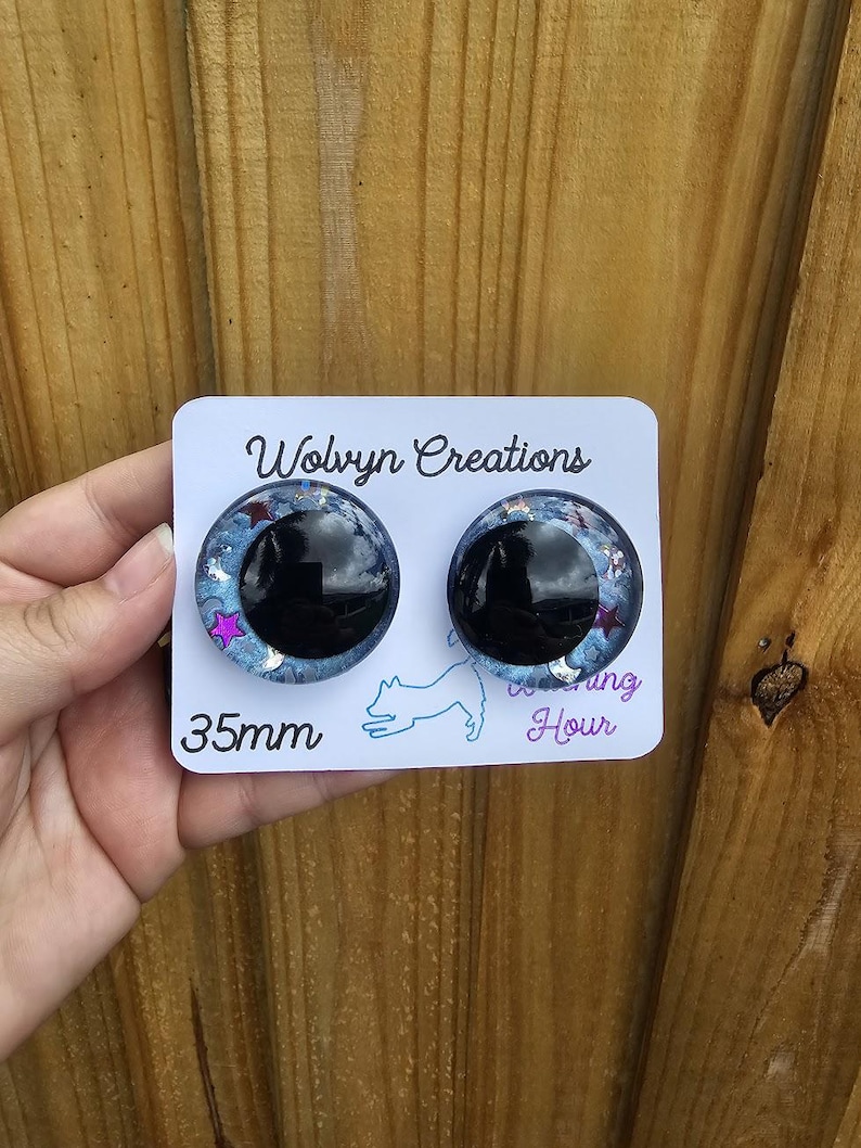 May include: A pair of 35mm round earrings with a black center and a blue, glittery outer ring. The earrings are on a white card with the text "Wolvyn Creations" and a dog silhouette. The earrings are held by a hand.