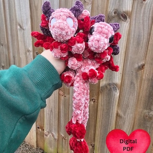 May include: Two plush axolotls in shades of pink, red, and purple. The handmade creatures have large, round eyes and detailed gills. The axolotls are held in a person's hand, with a wooden fence in the background. A red heart with the text "Digital PDF Pattern" is in the bottom right.