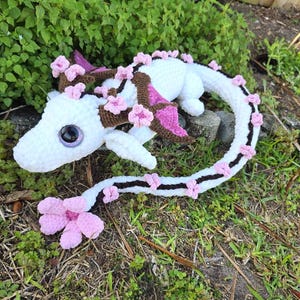 May include: A white crocheted dragon with pink and brown accents, adorned with pink flower embellishments. The dragon has large, dark eyes and is lying on a rock in a grassy area. The tail is long and detailed with pink flowers.