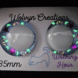 May include: Two round, glowing resin cabochons with a celestial theme. Each cabochon has a clear, domed center surrounded by a blue glitter border with stars and crescent moons. The text "Witching Hour" and "35mm" are visible.