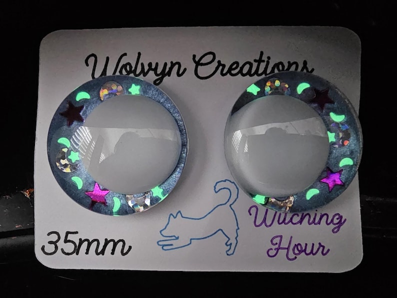 May include: Two round, glowing resin cabochons with a celestial theme. Each cabochon has a clear, domed center surrounded by a blue glitter border with stars and crescent moons. The text "Witching Hour" and "35mm" are visible.