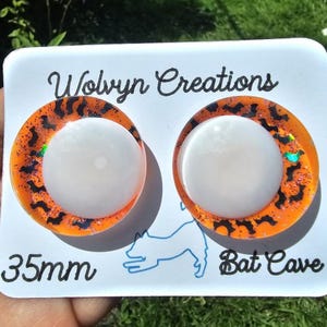May include: A pair of round earrings with a white center and an orange border with black bat silhouettes. The earrings are 35mm in diameter and are on a white card with the text "Wolvyn Creations" and "Bat Cave".