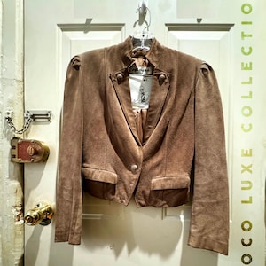 Puede incluir: Chaqueta de ante marrón con un solo botón, hombreras y dos bolsillos delanteros. La chaqueta está colgada de una percha transparente contra una puerta blanca. El texto "COCO LUXE COLLECTION" es visible en el lado derecho.