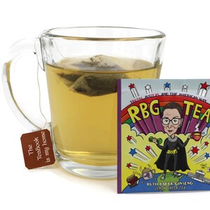 May include: A glass mug filled with green tea with a tea bag that says "The TeaBook is my home".  A tea bag packet with a cartoon image of Ruth Bader Ginsburg in a superhero costume with the text "Truth, Justice, and the American Way RBG TEA Ruth Bader Ginseng Lemon Green Tea"