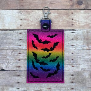 Op de afbeelding: Een regenboog glitter sleutelhanger met een zwart silhouet van vleermuizen. De sleutelhanger heeft een zilveren metalen ring en een zwarte knop.
