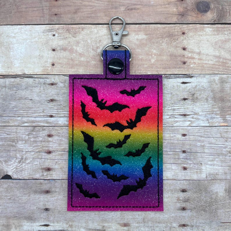 Op de afbeelding: Een regenboog glitter sleutelhanger met een zwart silhouet van vleermuizen. De sleutelhanger heeft een zilveren metalen ring en een zwarte knop.