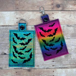 Op de afbeelding: Twee sleutelhangers met een regenboog en iriserende achtergrond met zwarte vleermuis silhouetten.