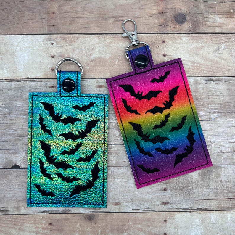 Op de afbeelding: Twee sleutelhangers met een regenboog en iriserende achtergrond met zwarte vleermuis silhouetten.