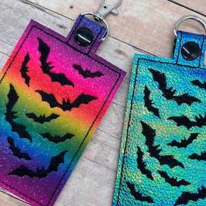 Op de afbeelding: Twee sleutelhangers met zwarte vleermuizen op een kleurrijke achtergrond. De ene sleutelhanger heeft een regenboogachtergrond en de andere een blauw-groene, iriserende achtergrond.