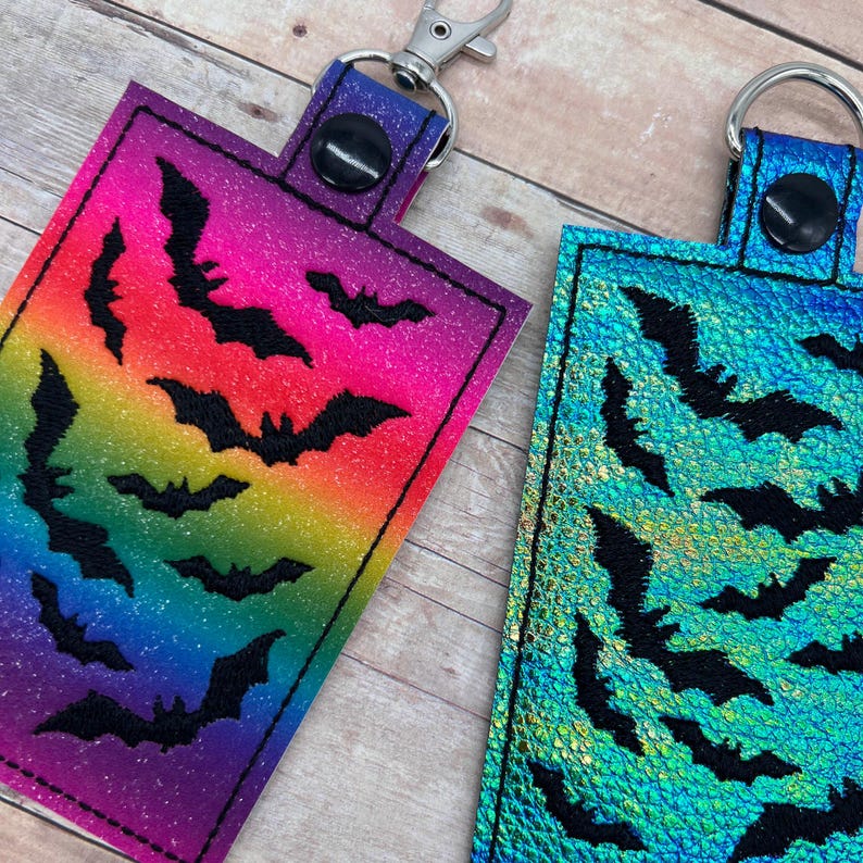 Op de afbeelding: Twee sleutelhangers met zwarte vleermuizen op een kleurrijke achtergrond. De ene sleutelhanger heeft een regenboogachtergrond en de andere een blauw-groene, iriserende achtergrond.