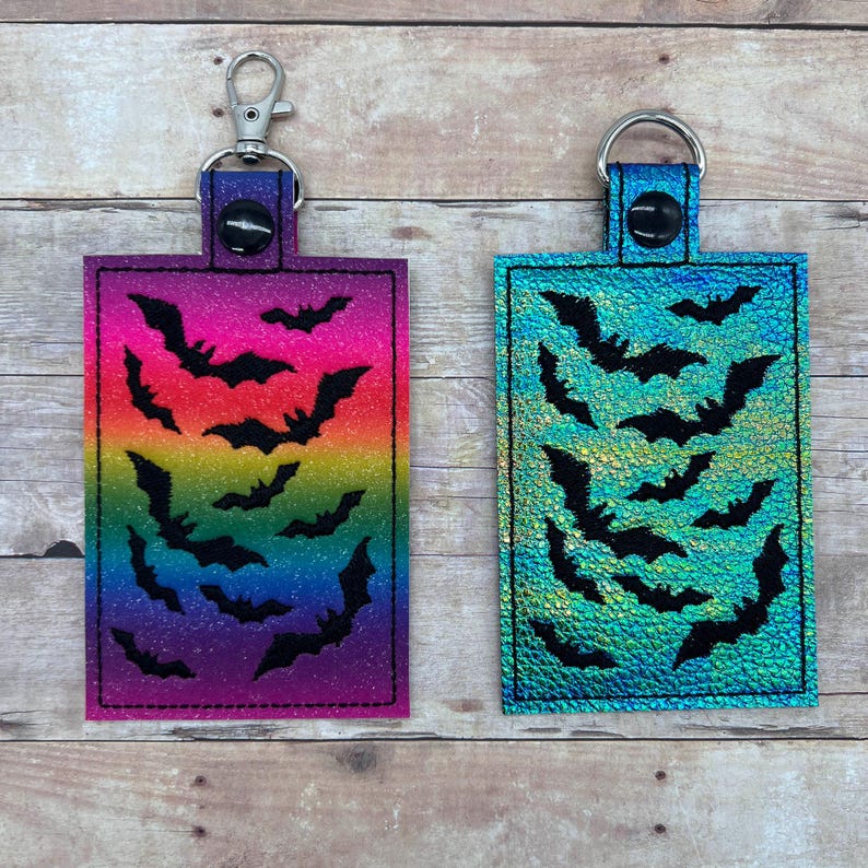 Op de afbeelding: Twee sleutelhangers met zwarte vleermuizen op een glinsterende achtergrond. De ene sleutelhanger heeft een regenboogkleurige glitterachtergrond en de andere een turquoise glitterachtergrond.