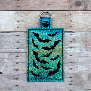 Op de afbeelding: Een turquoise blauwe sleutelhanger met een zilveren ring en een ontwerp van zwarte vleermuizen op een glinsterende achtergrond.