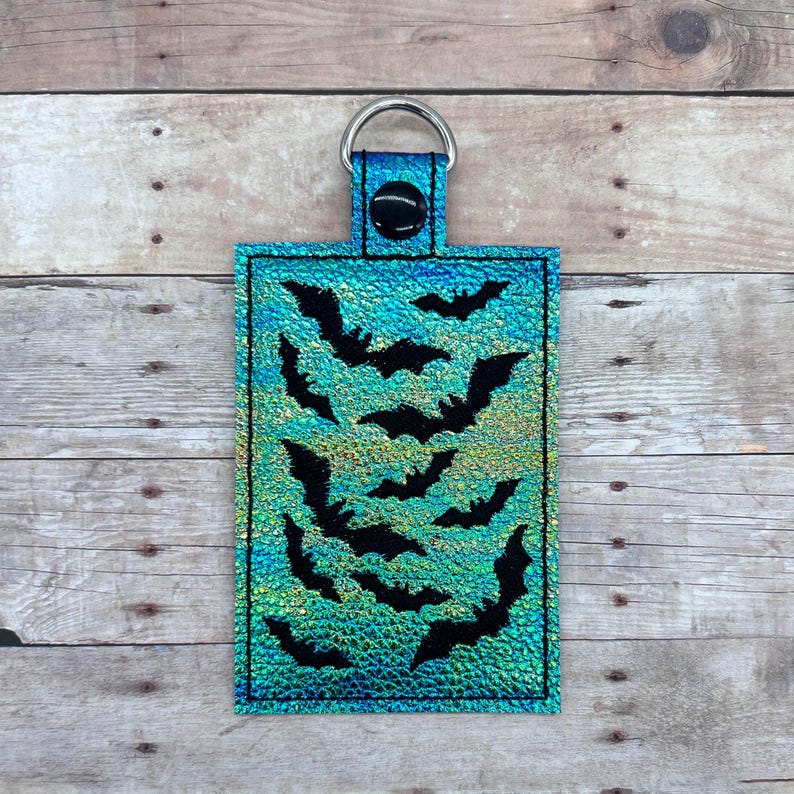 Op de afbeelding: Een turquoise blauwe sleutelhanger met een zilveren ring en een ontwerp van zwarte vleermuizen op een glinsterende achtergrond.