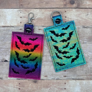 Op de afbeelding: Twee sleutelhangers met een regenboog en turquoise iriserende achtergrond en zwarte vleermuis silhouetten. De sleutelhangers hebben een zilveren metalen ring en een zwarte drukknoop sluiting.