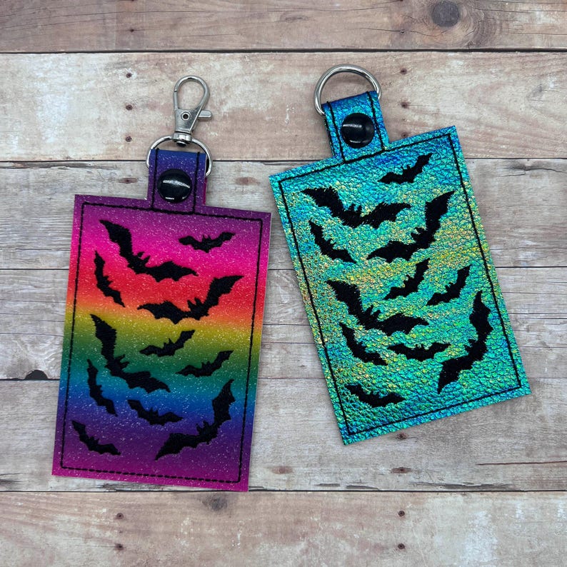 Op de afbeelding: Twee sleutelhangers met een regenboog en turquoise iriserende achtergrond en zwarte vleermuis silhouetten. De sleutelhangers hebben een zilveren metalen ring en een zwarte drukknoop sluiting.