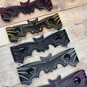 May include: Five different colored leather bat-shaped mask extenders with black buttons. The colors are red, yellow, black, gray, and purple.